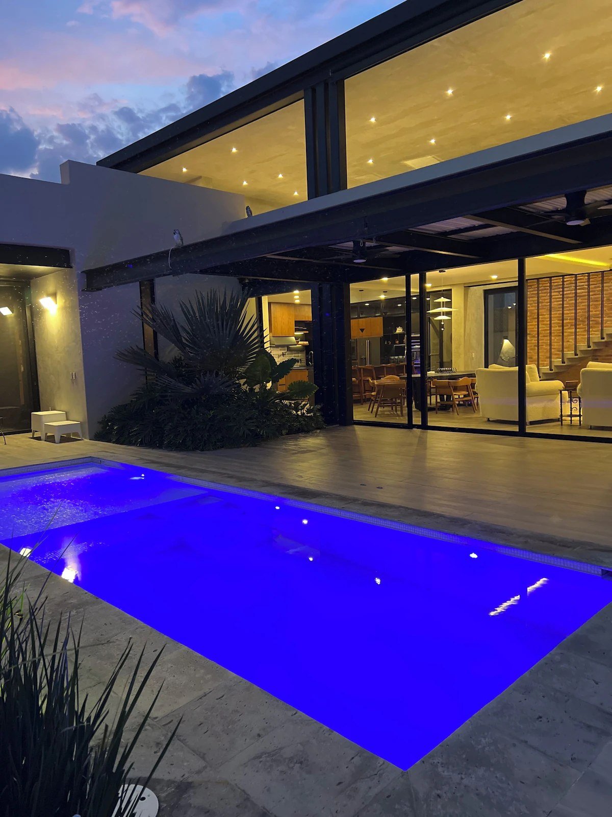 The house at dusk, pool lit, glass walls open to the living room and kitchen